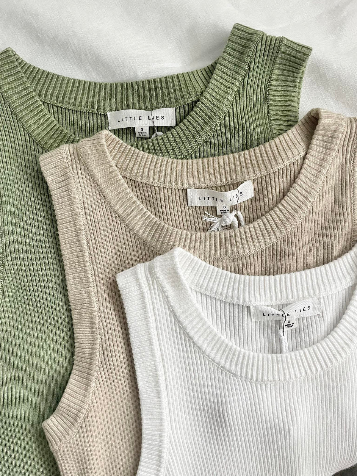 Three ribbed knit tank tops in sage green, beige, and white, arranged in an overlapping flat-lay. Each top has a white neck label reading 'LITTLE LIES'.