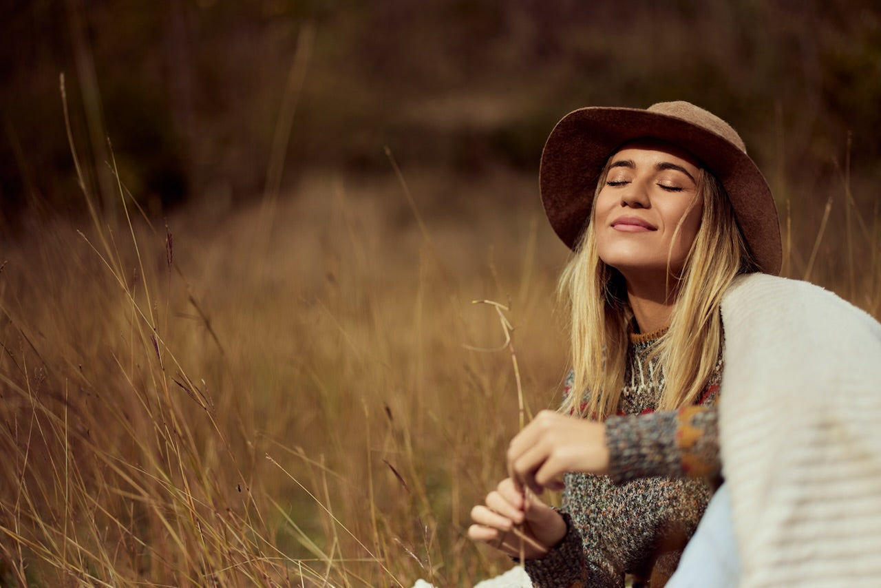 Woman with eyes closed, smiling, enjoying Autumn sunshine. She wears a brown hat, with blonde hair out wearing grey knit and beige shawl in outdoor grassy area.