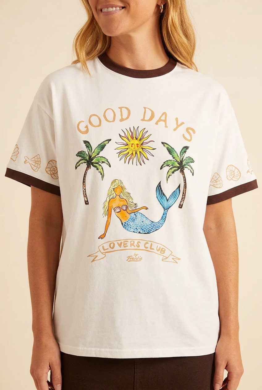 A woman wearing an oversized white graphic t-shirt with brown trim on the collar and sleeve cuffs. The shirt features a colorful graphic of a mermaid, palm trees, a sun, and the text 'GOOD DAYS LOVERS CLUB' on the front.