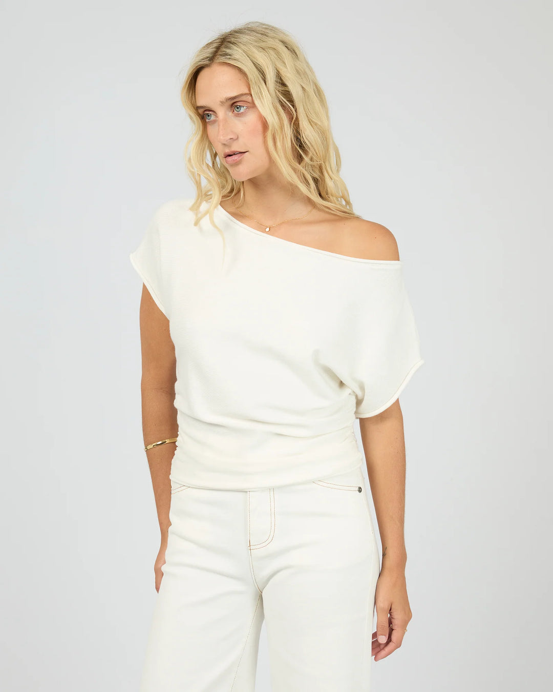 A blonde woman wearing a cream off-the-shoulder knit top and matching cream pants, photographed against a light gray studio background.