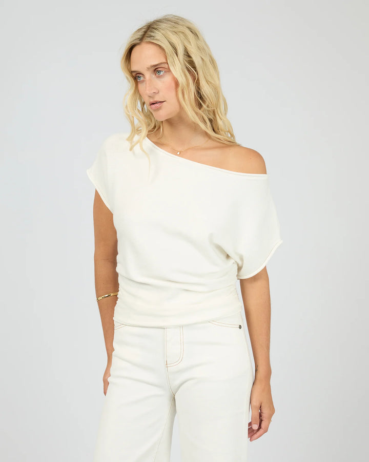 A blonde woman wearing a cream off-the-shoulder knit top and matching cream pants, photographed against a light gray studio background.