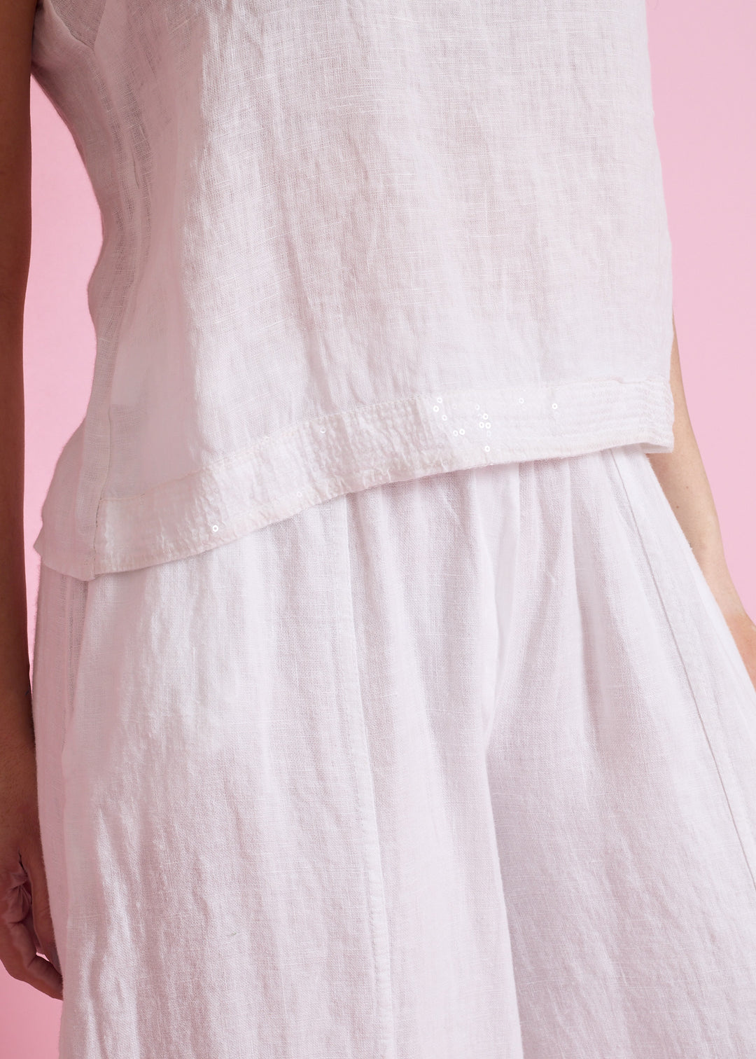 A close-up of a model wearing a light pink linen top and matching wide-leg skirt against a solid pink background.