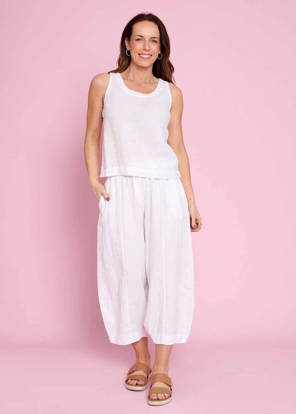A woman modeling a white sleeveless top and matching white wide-leg cropped pants against a solid pink studio background.
