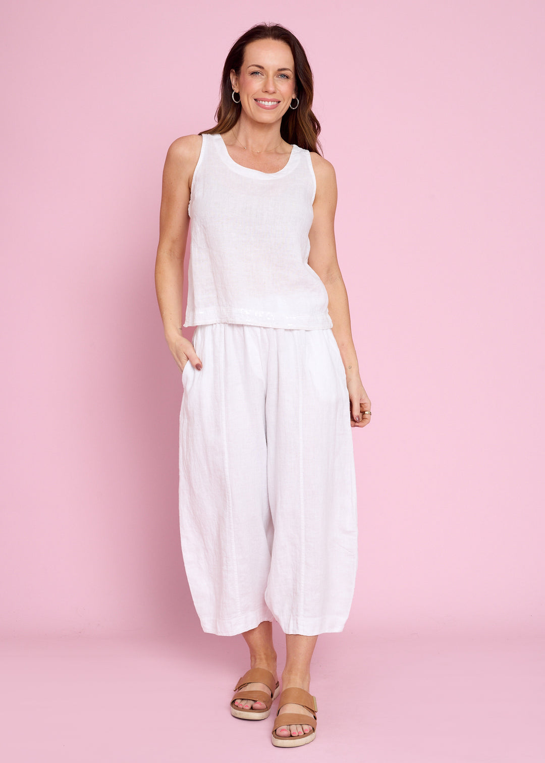 A woman modeling a white sleeveless top and matching white wide-leg cropped pants against a solid pink studio background.