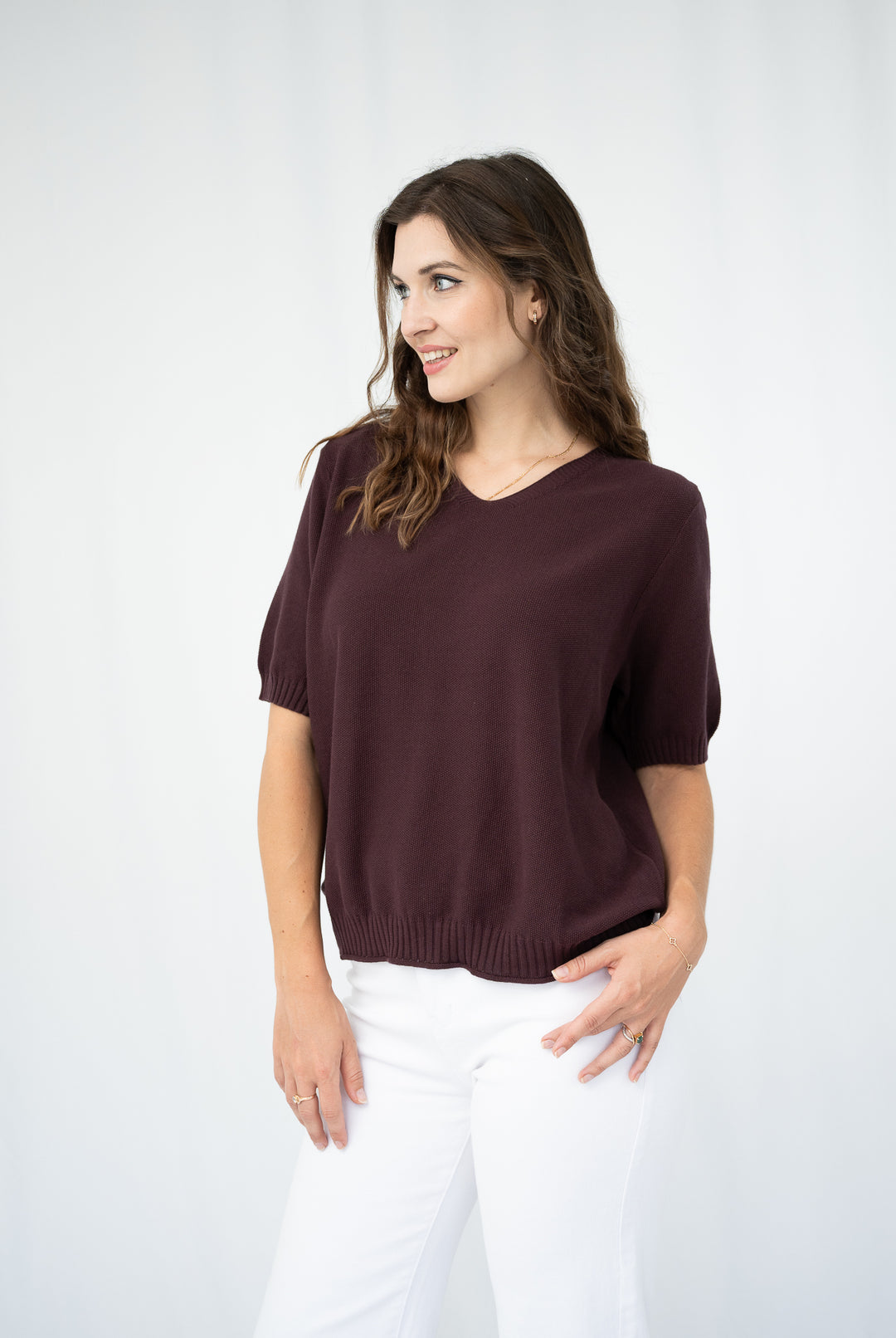 A woman wearing a short-sleeve knit sweater in deep burgundy with white pants, standing against a plain light gray studio background.
