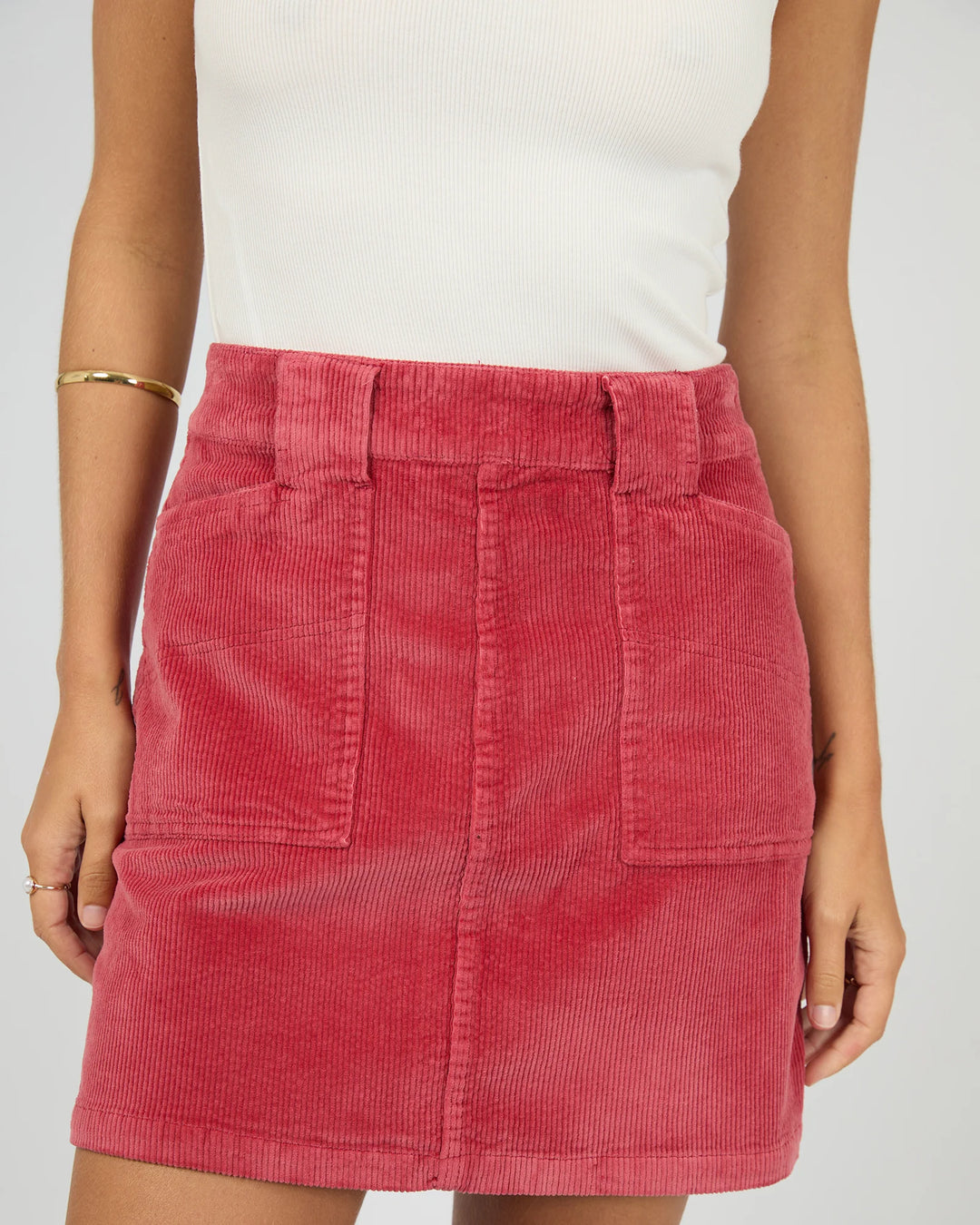 A model wearing a pink corduroy mini skirt with front patch pockets and a matching corduroy belt, paired with a white ribbed tank top against a light gray background.