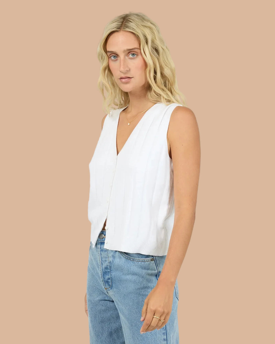 A blonde woman wearing a white sleeveless V-neck top and light blue jeans, standing against a light gray studio background.