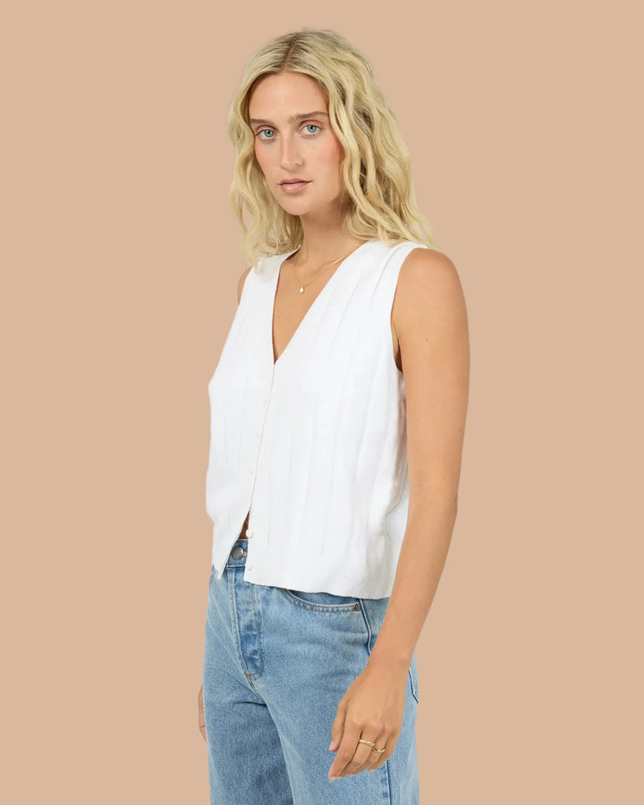 A blonde woman wearing a white sleeveless V-neck top and light blue jeans, standing against a light gray studio background.