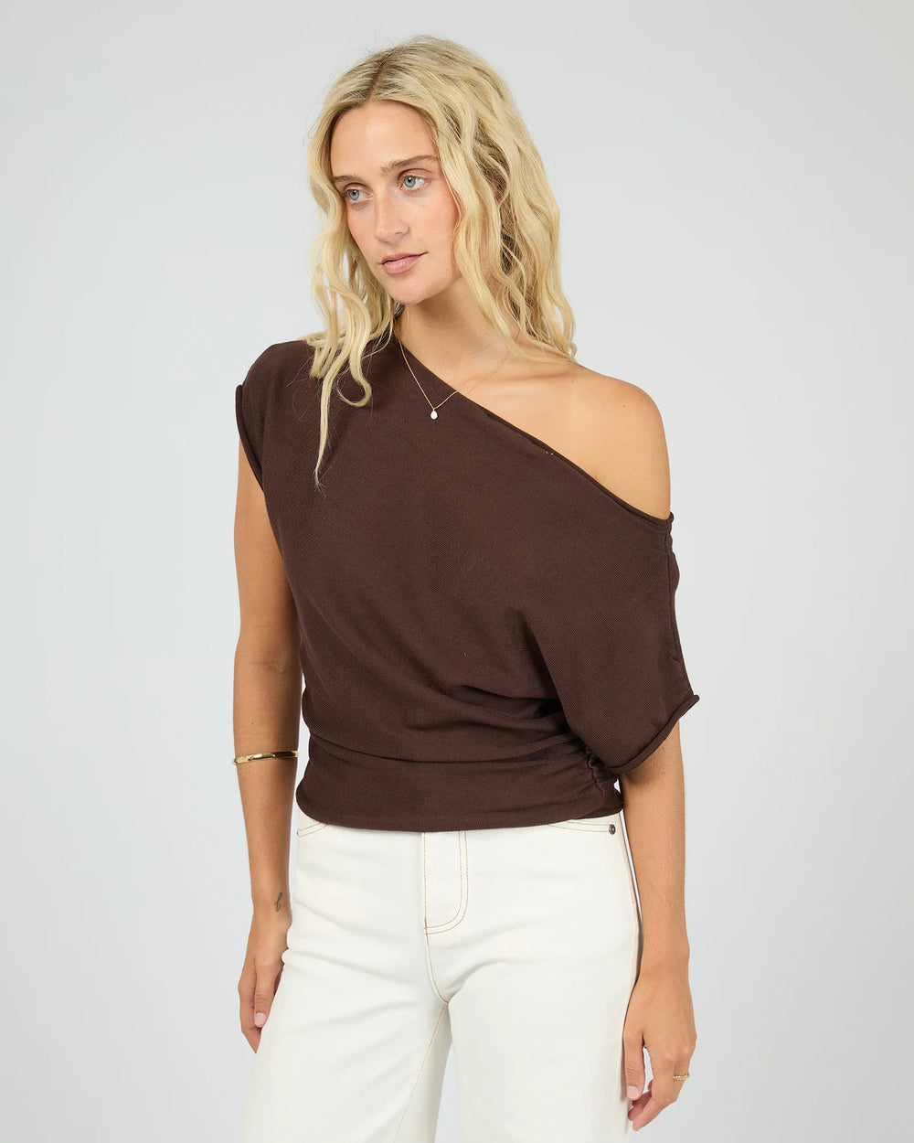 Woman wearing a brown one-shoulder top and white pants on a plain background