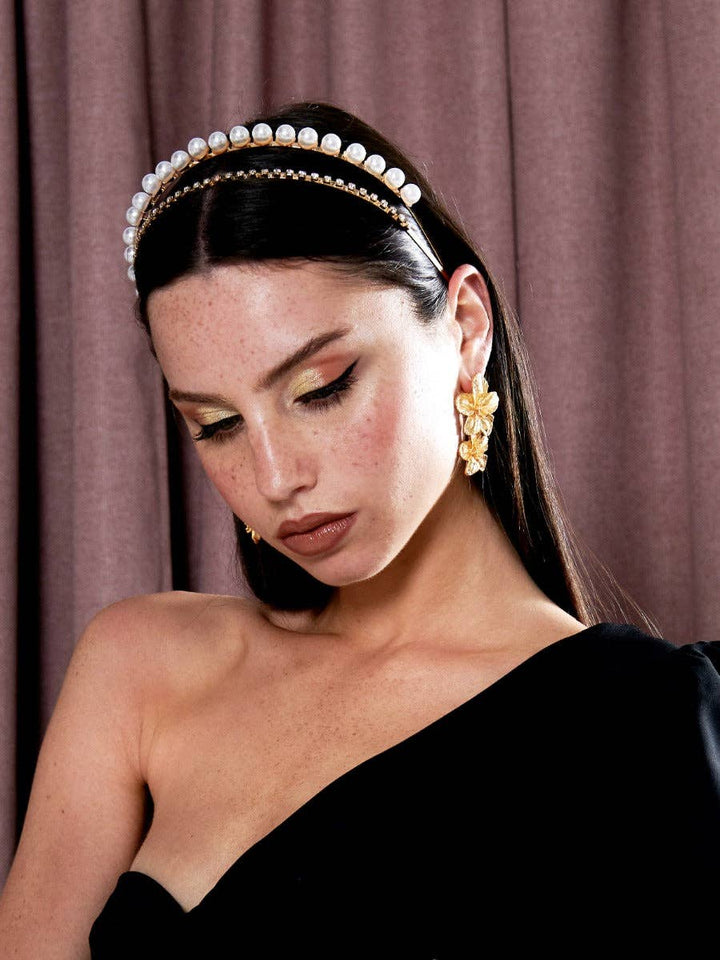 Woman wearing a pearl headband and gold earrings against a brown curtain background