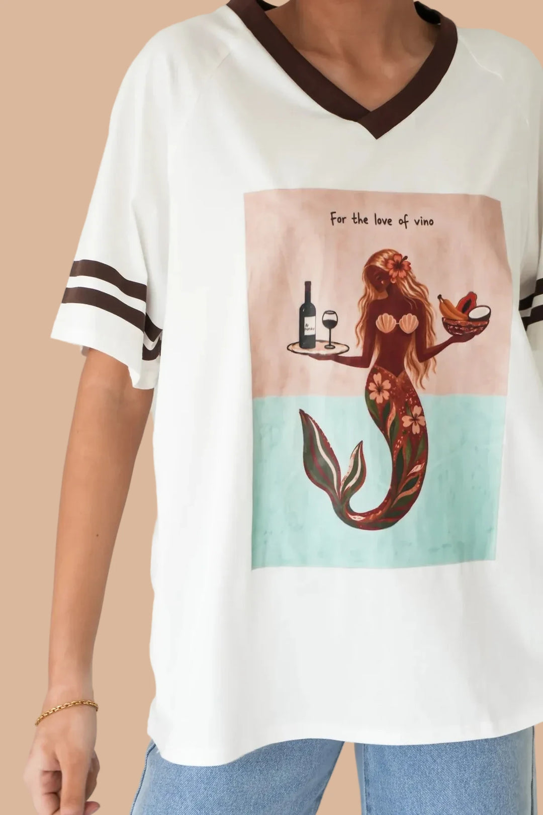 A woman wearing a white V-neck t-shirt with dark brown trim on the collar and sleeve cuffs, featuring a graphic print of a mermaid serving a wine bottle and glass on a tray, with the text &