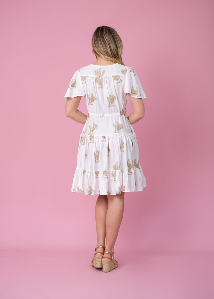 A woman stands with her back to the camera wearing a white short-sleeve dress with a beige leaf pattern, paired with tan wedge sandals, set against a solid pink studio background.