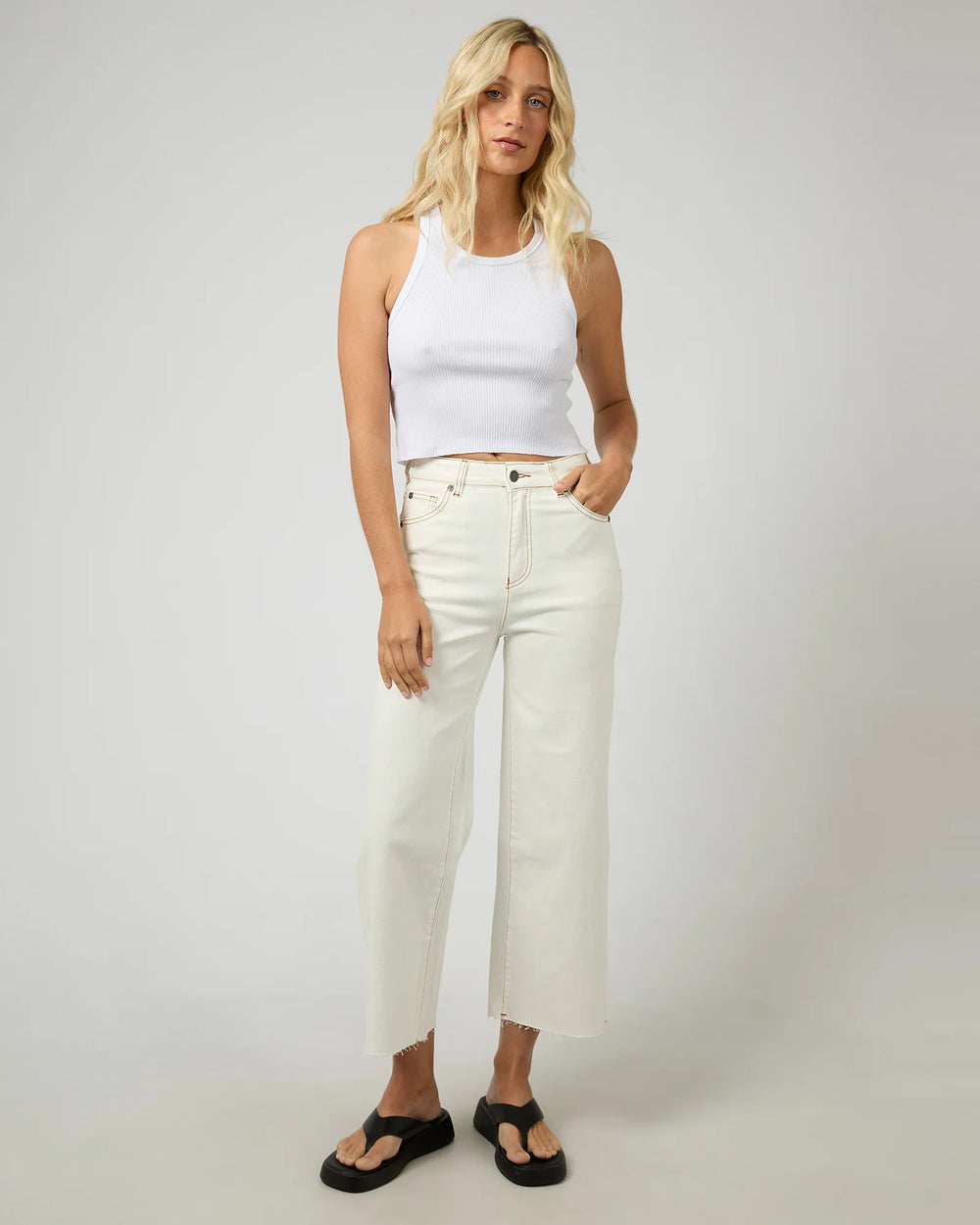 Woman wearing a white tank top and white jeans against a plain background