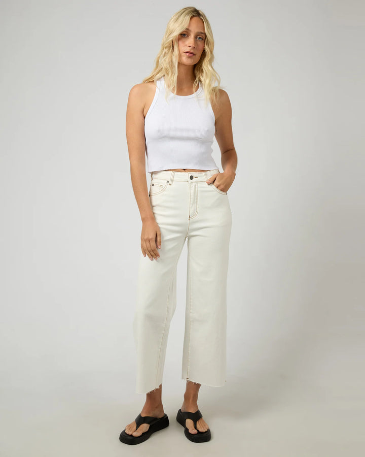 Woman wearing a white tank top and white jeans against a plain background