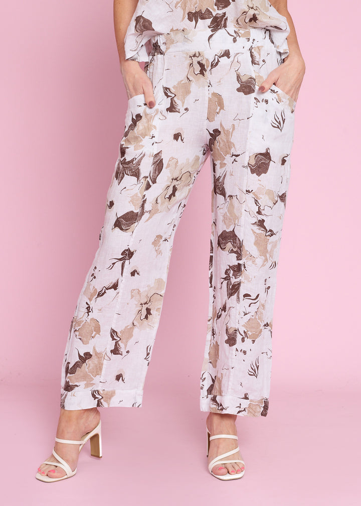 A model wearing white wide-leg trousers with a brown and beige floral print, paired with white strappy high-heeled sandals, set against a solid pink studio background.