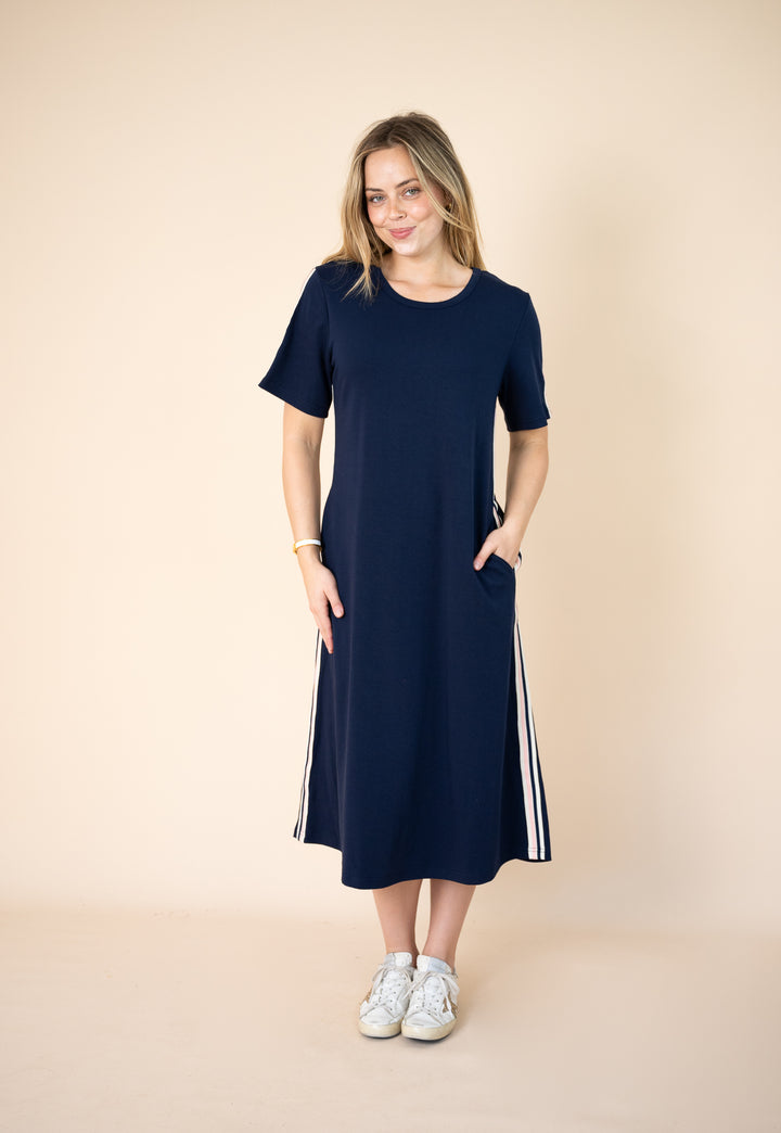 A woman modeling a navy blue short-sleeve midi dress with white side stripes, standing against a solid beige studio background.