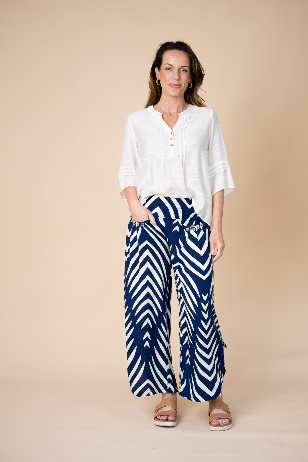 A woman modeling a white button-up blouse and navy blue wide-leg pants with a white chevron pattern, standing against a solid beige studio background.