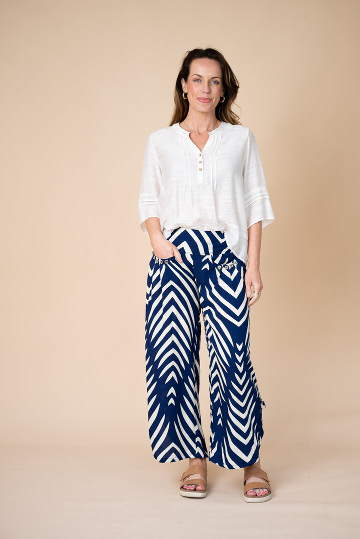 A woman modeling a white button-up blouse and navy blue wide-leg pants with a white chevron pattern, standing against a solid beige studio background.