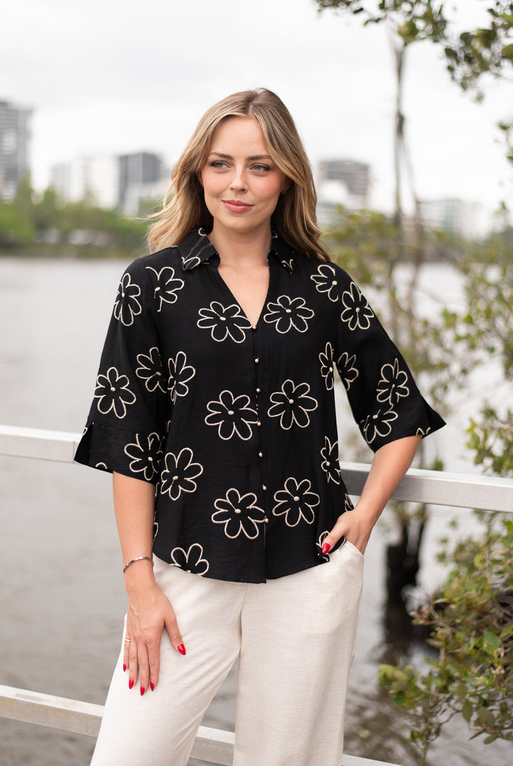 Woman wearing a black floral blouse and white pants standing by a waterfront with trees and buildings in the background.