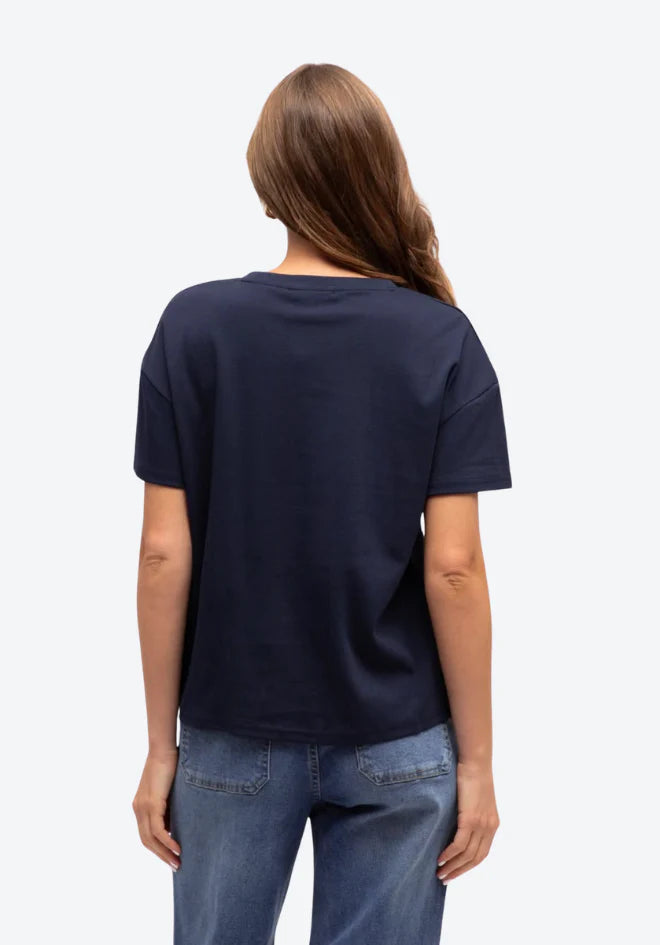 A woman viewed from the back wearing a dark navy blue short-sleeve t-shirt and blue jeans against a light gray background.