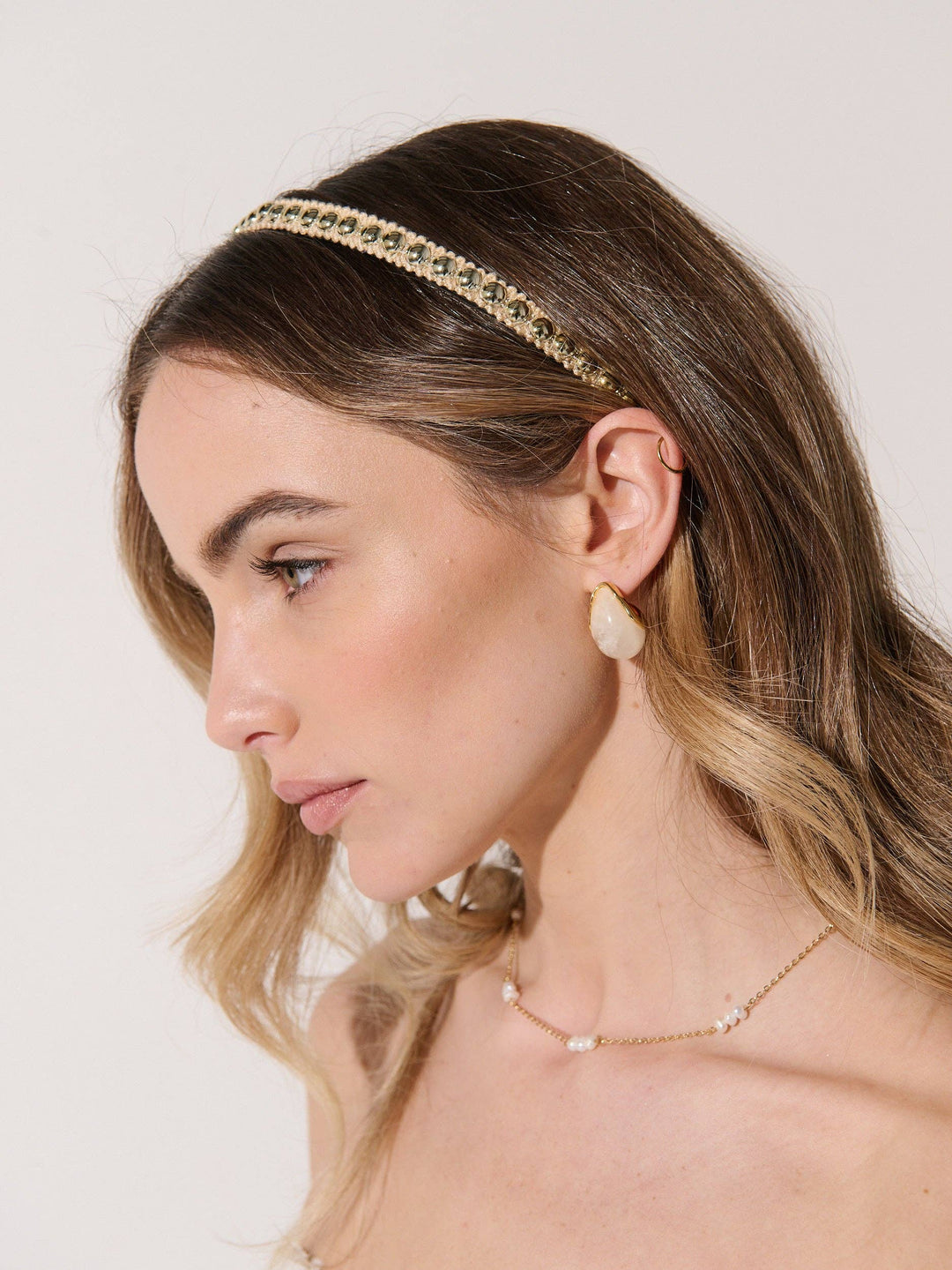 A woman shown in profile wearing a gold chain headband embellished with small clear stones, along with matching gold jewelry against a plain light background.
