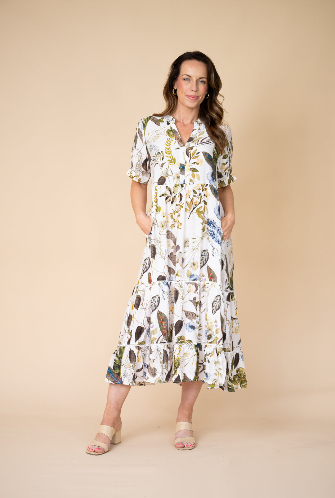 A woman modeling a white midi dress with a multicolor botanical and abstract leaf print, featuring short puff sleeves, a V-neckline, and a tiered ruffle hem, set against a solid beige studio background.