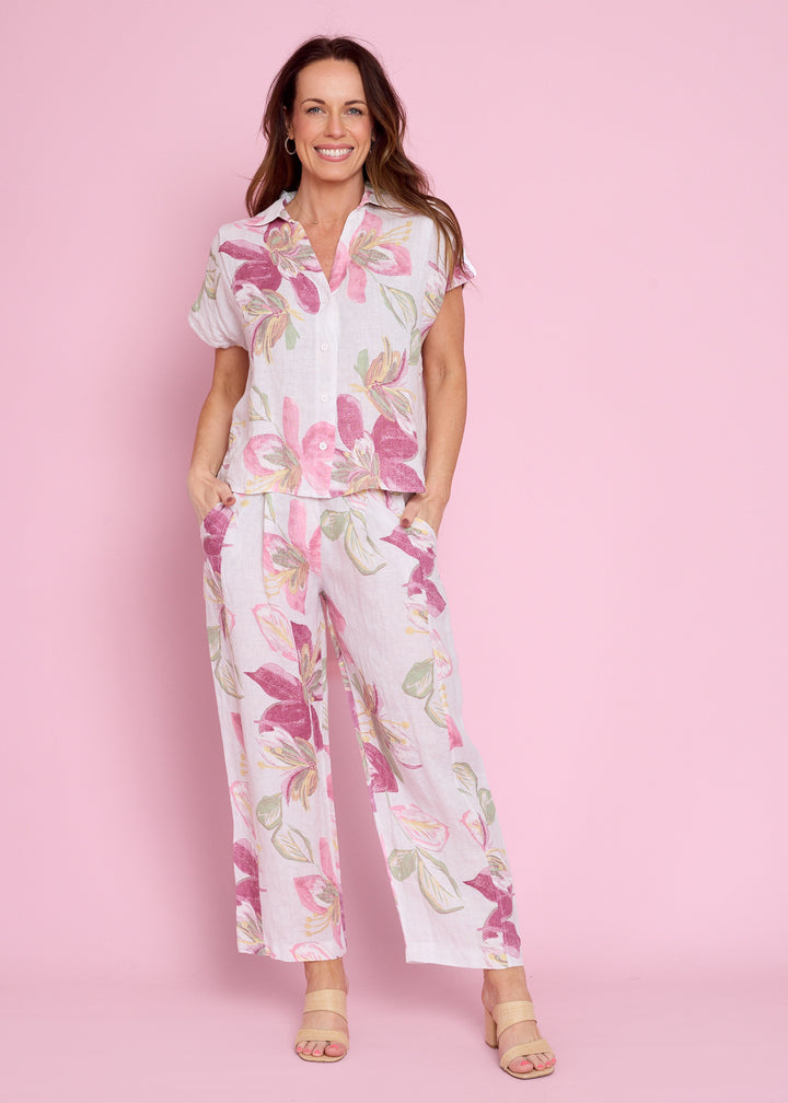 A woman modeling a matching short-sleeve button-up shirt and wide-leg pants set in a white fabric with a pink and green floral print, set against a solid pink background.