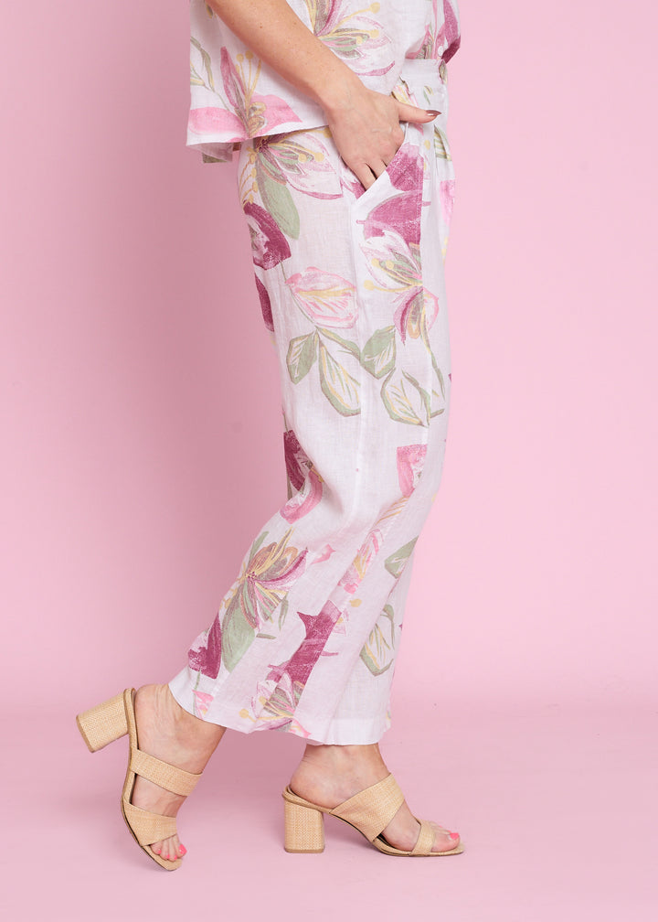 A person wearing white cropped pants with a pink, green, and yellow floral print, paired with beige block-heel sandals, set against a solid pink background.