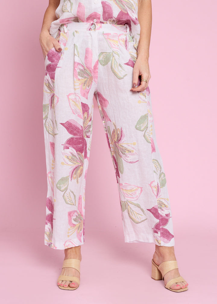 A model wearing high-waisted, wide-leg cropped pants with a pink, green, and beige floral print on a white base, paired with beige block-heel sandals against a solid pink background.
