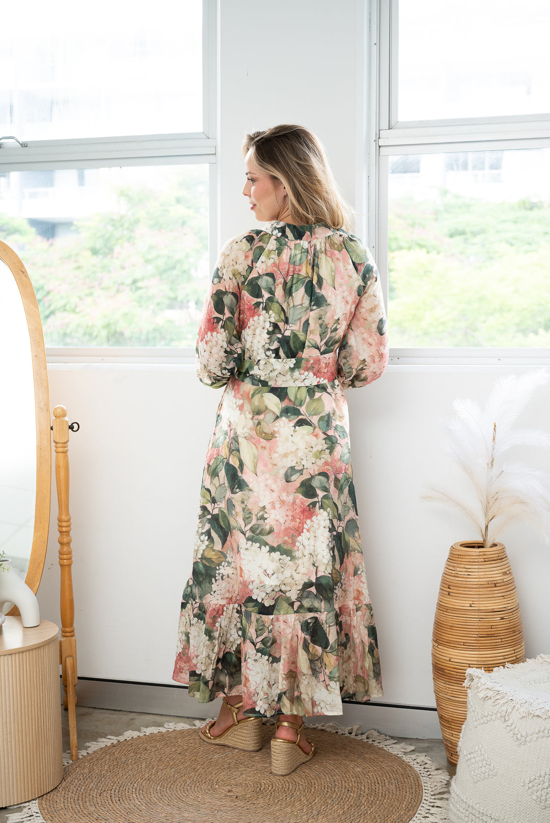 Woman in a long sleeve, long floral green, pink and white  dress standing in a room with large windows and decorative elements.