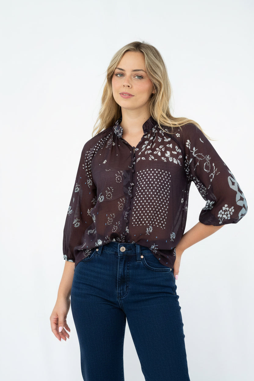 A woman modeling a sheer dark brown button-up blouse with a white floral and polka dot pattern, paired with light blue jeans, against a plain white studio background.