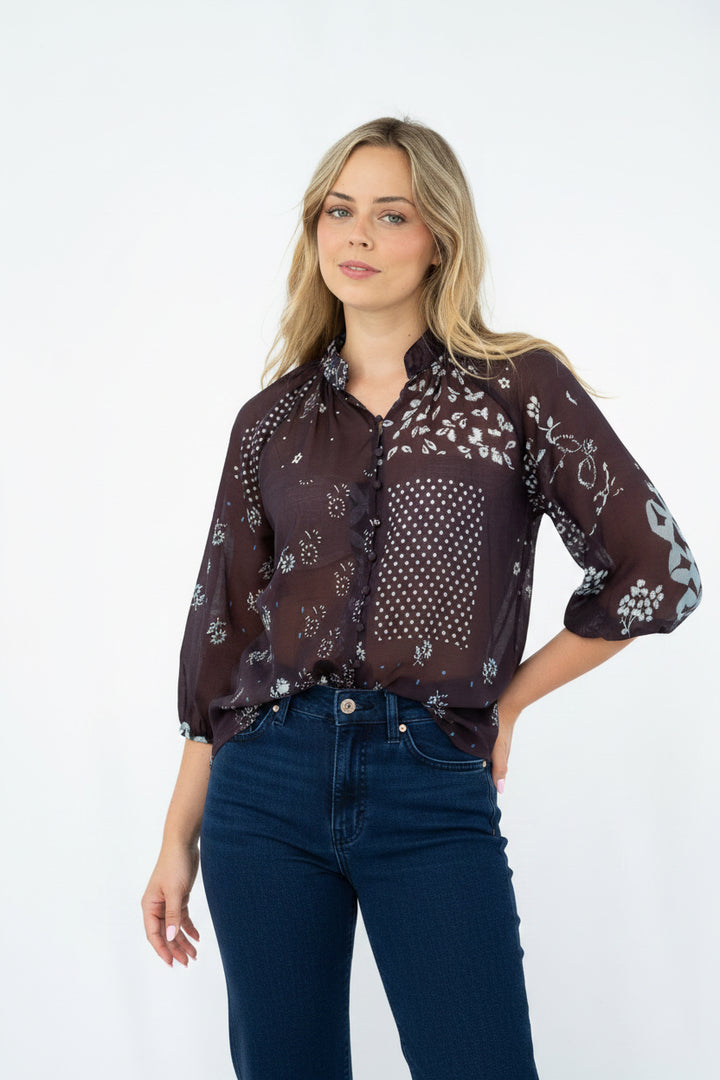 A woman modeling a sheer dark brown button-up blouse with a white floral and polka dot pattern, paired with light blue jeans, against a plain white studio background.