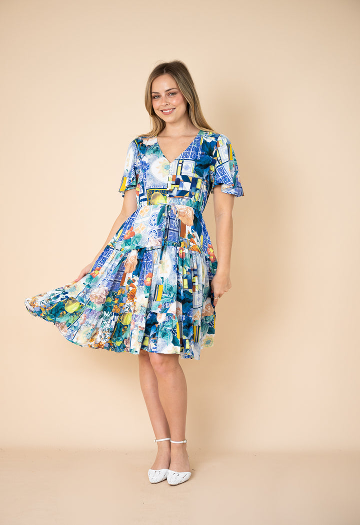 A woman modeling a short-sleeve V-neck A-line dress with a colorful abstract patchwork print in shades of blue, green, yellow, and white, set against a solid beige studio background.