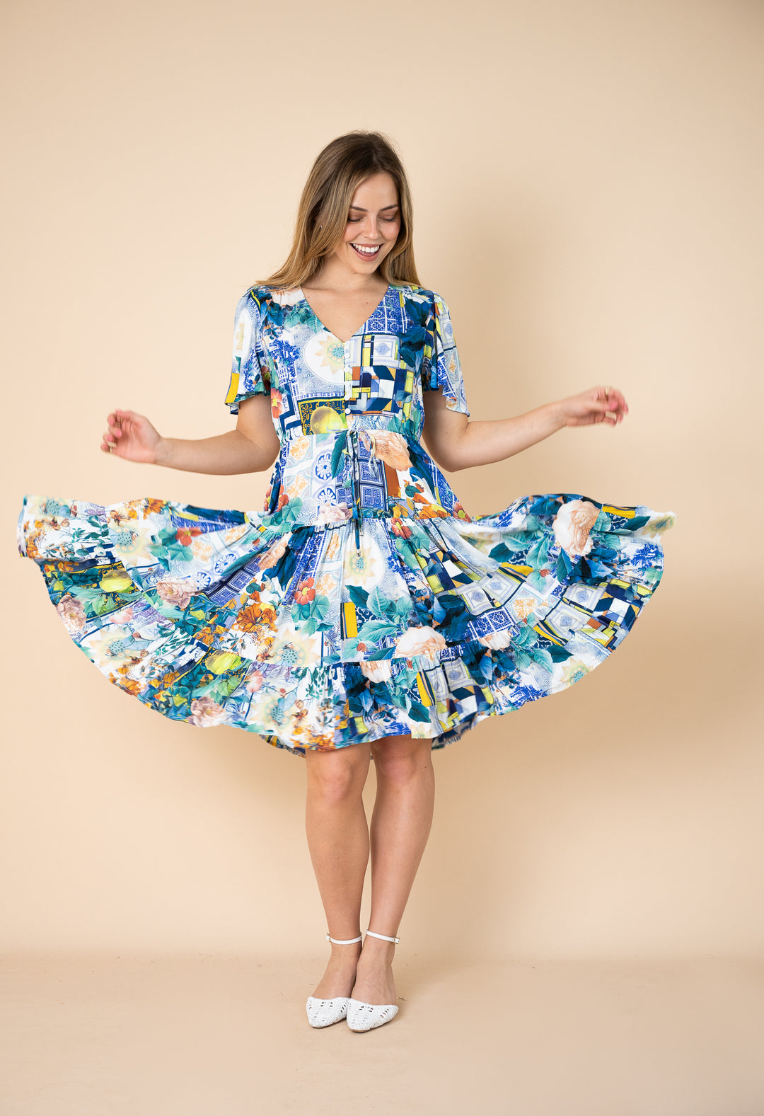 A woman wearing a short-sleeve V-neck dress with a colorful abstract patchwork print in blue, white, yellow, and green, twirling to show off the flared skirt against a solid beige studio background.