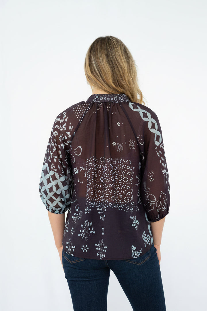 A woman viewed from the back wearing a sheer dark brown bandana-print blouse with white patterns and light blue jeans, set against a plain white studio background.