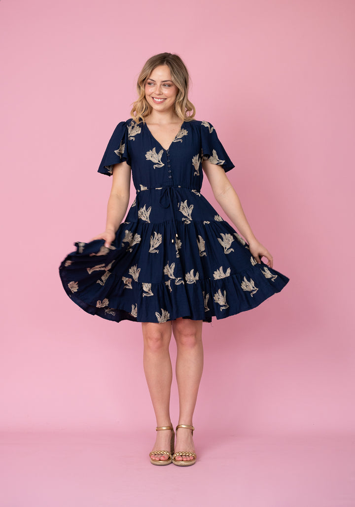 A woman modeling a navy blue short-sleeve V-neck dress with a beige floral print, standing against a solid pink studio background.