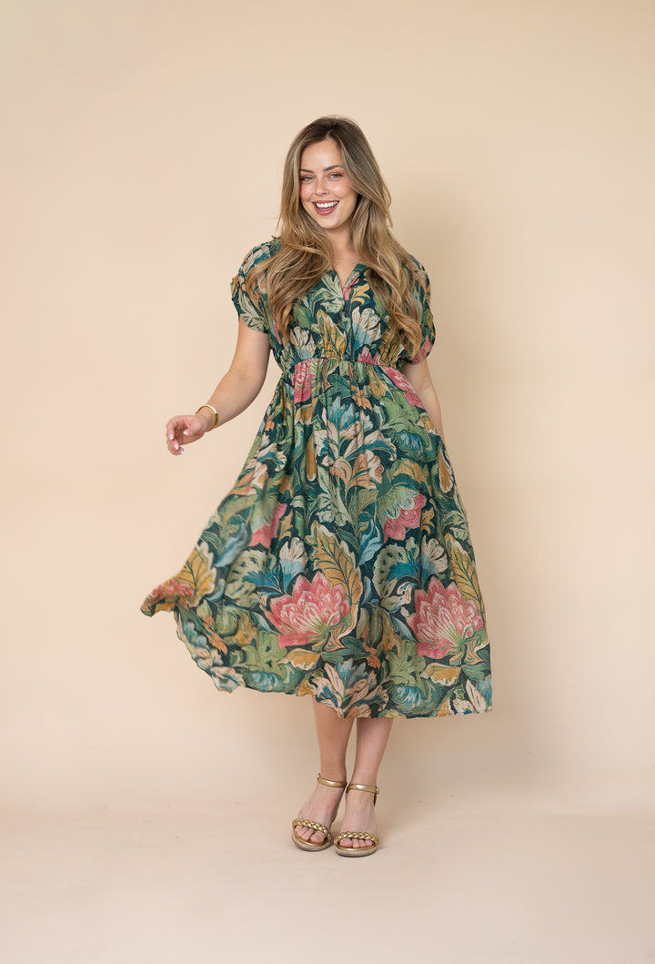 A woman modeling a short-sleeve midi dress with a colorful floral print in green, pink, and yellow, set against a solid beige studio background.