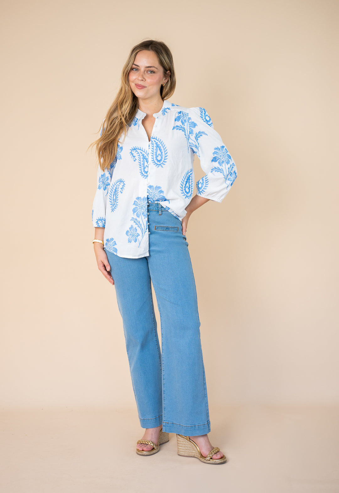 A woman modeling a white long-sleeve button-up blouse with a blue paisley pattern, paired with light blue wide-leg jeans and beige wedge sandals, against a solid beige studio background.