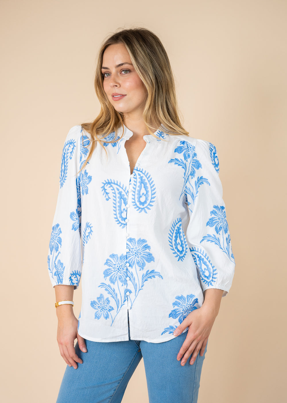 A woman wearing a white long-sleeve button-up blouse with a blue paisley and floral print, paired with blue jeans, against a solid beige background.