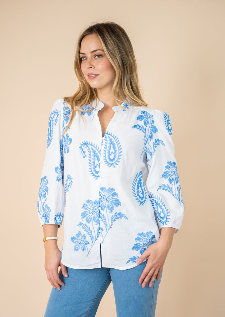 A woman wearing a white long-sleeve button-up blouse with a blue paisley and floral print, paired with blue jeans, against a solid beige background.