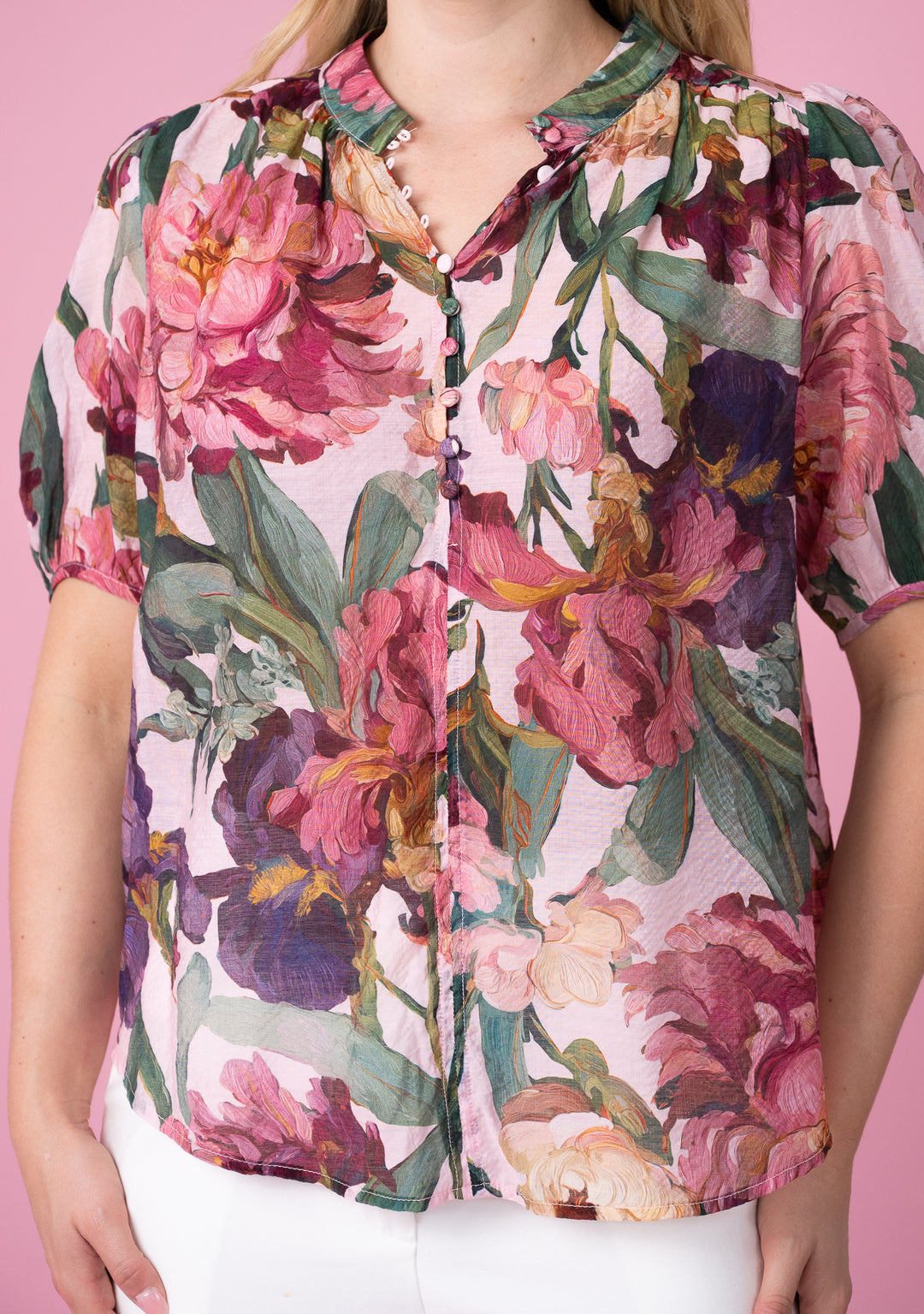 A woman wearing a short-sleeve button-up blouse with a vibrant pink, purple, and green floral print, paired with white pants, set against a solid pink background.