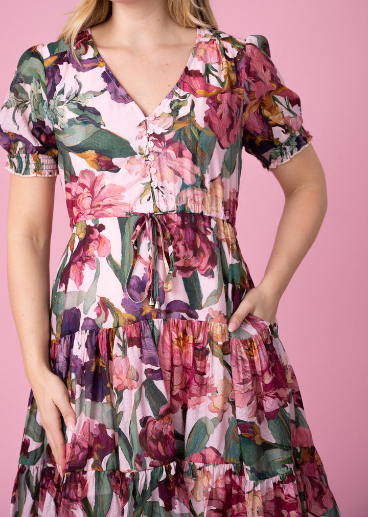 A woman wearing a short-sleeve tiered dress with a pink, purple, and green floral print, standing against a solid pink background.