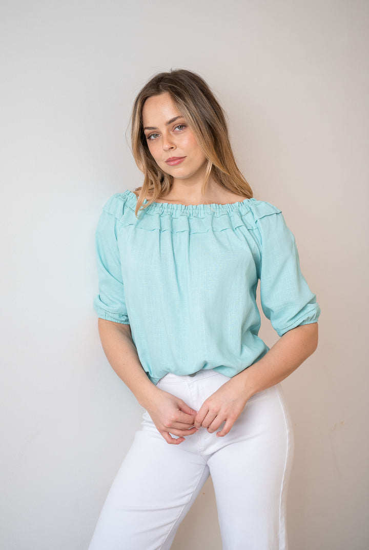 A woman modeling a light blue off-the-shoulder blouse with gathered puff sleeves, paired with white pants, standing against a plain light beige wall.