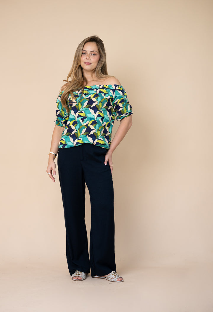 A woman modeling an off-the-shoulder short-sleeve top with a colorful abstract leaf pattern in teal, green, yellow, and black, paired with dark navy wide-leg pants, against a solid beige studio background.