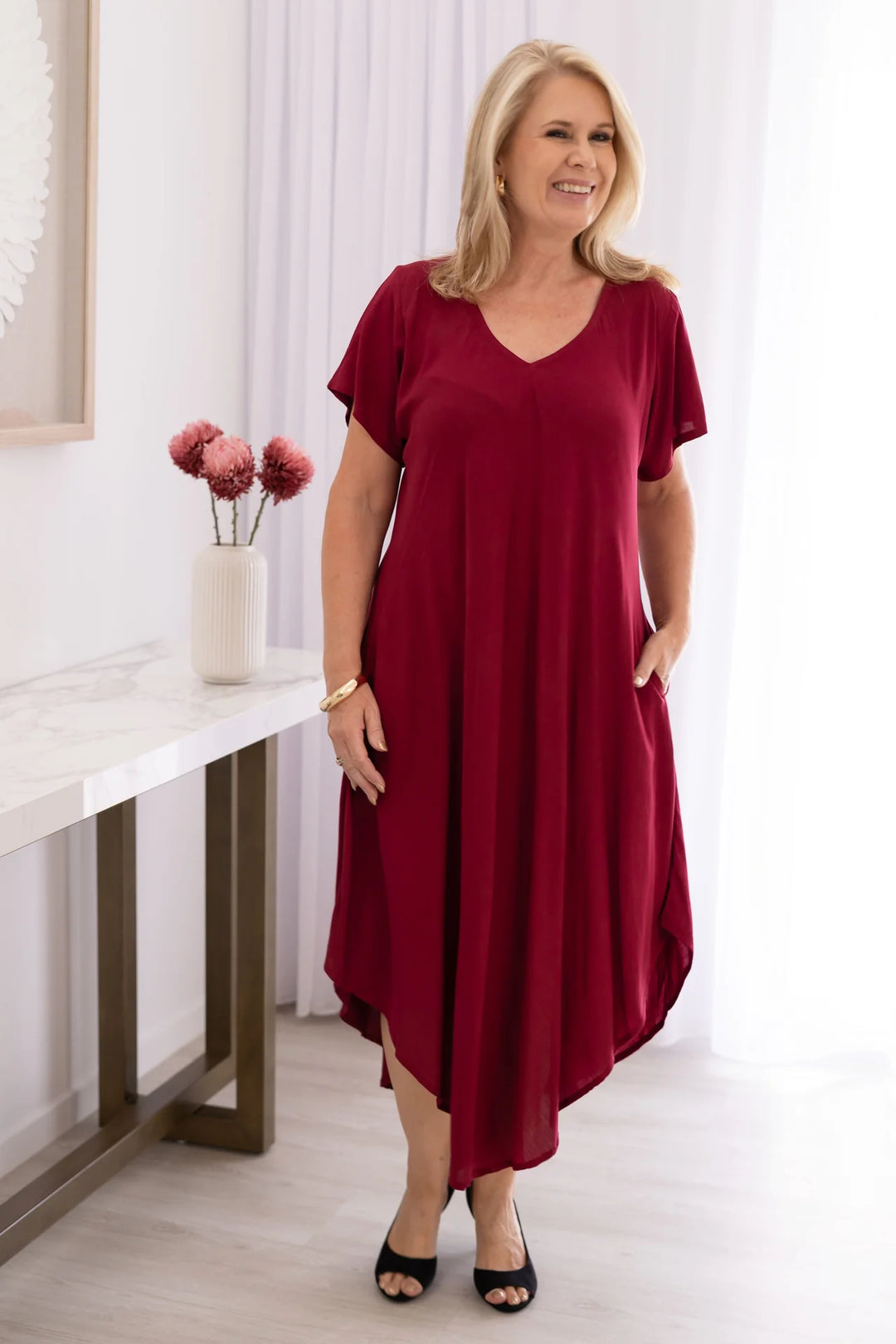 Woman wearing burgundy short sleeve dress smiling and standing beside hallway table in brightly lit room.