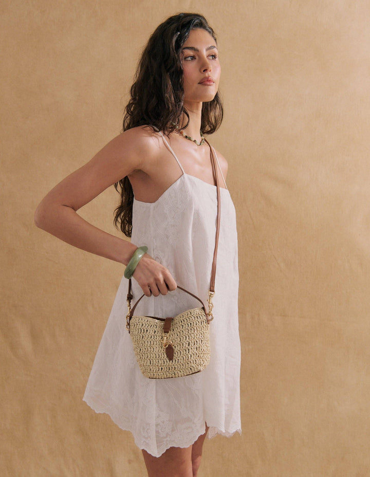 A woman wearing a white sleeveless dress stands against a warm beige textured backdrop, holding a small woven straw shoulder bag with tan straps.