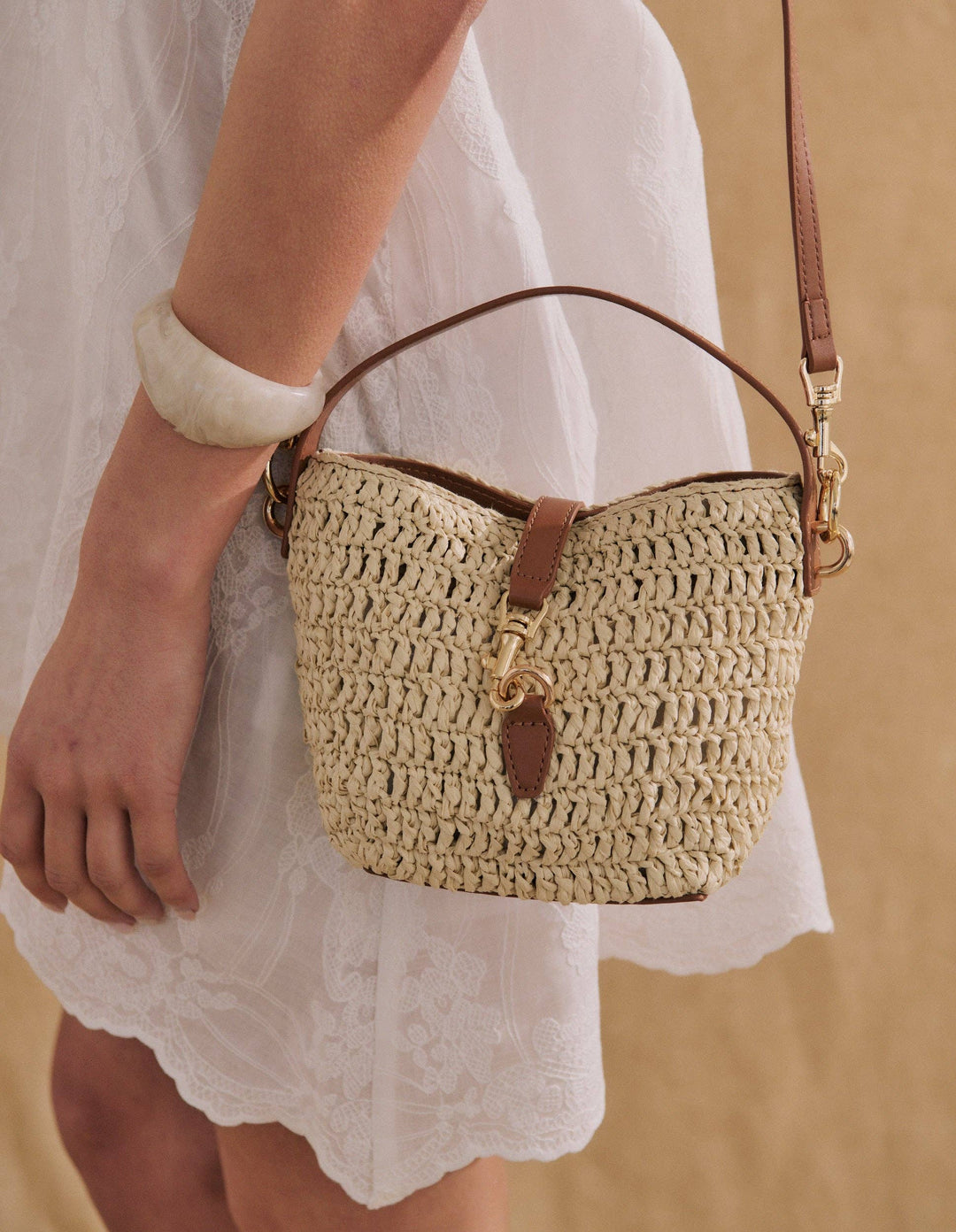 A woven straw bucket bag with brown leather trim and a gold-tone clasp, carried on a model wearing a white lace dress.