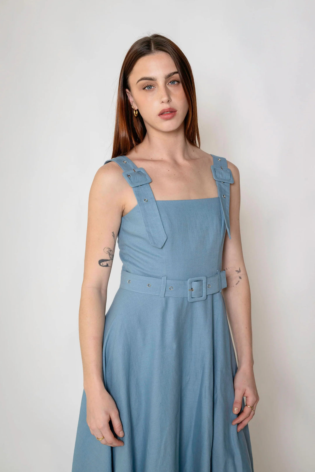 A woman modeling a light blue sleeveless dress with structured shoulder straps and a matching belt with a square buckle, photographed against a plain light gray studio background.