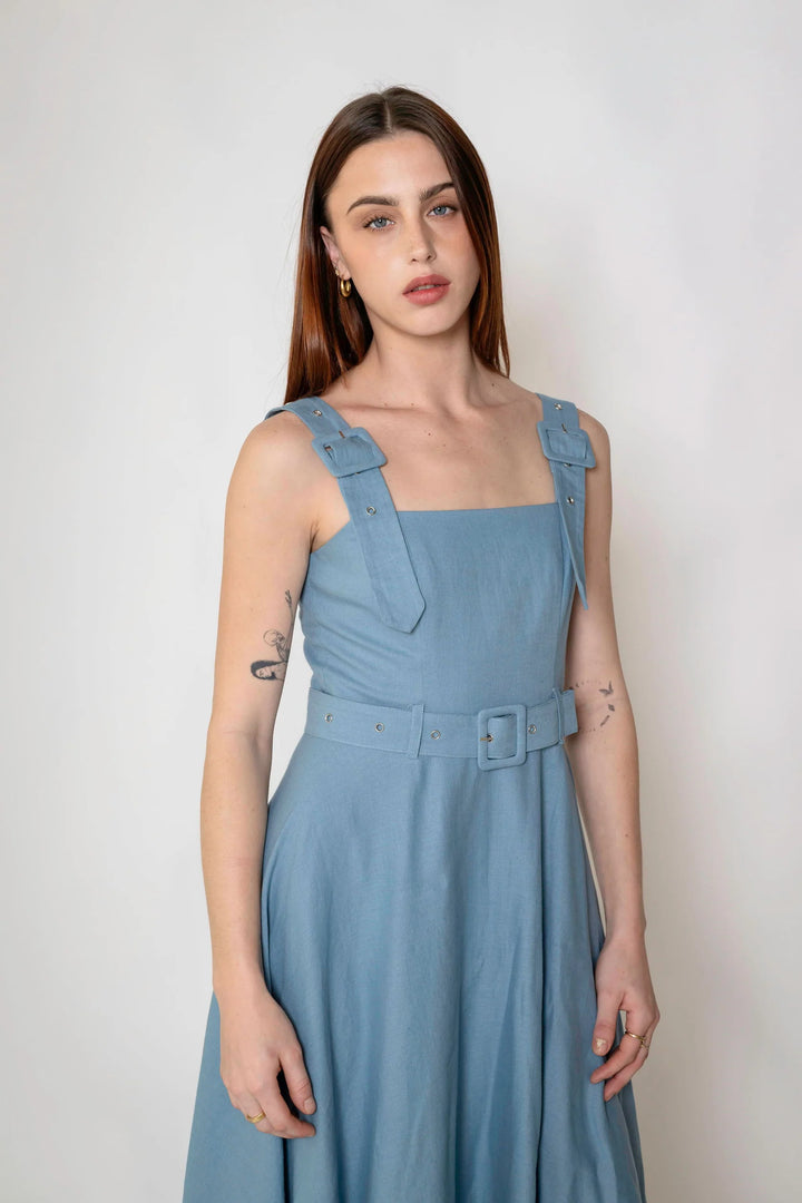 A woman modeling a light blue sleeveless dress with structured shoulder straps and a matching belt with a square buckle, photographed against a plain light gray studio background.