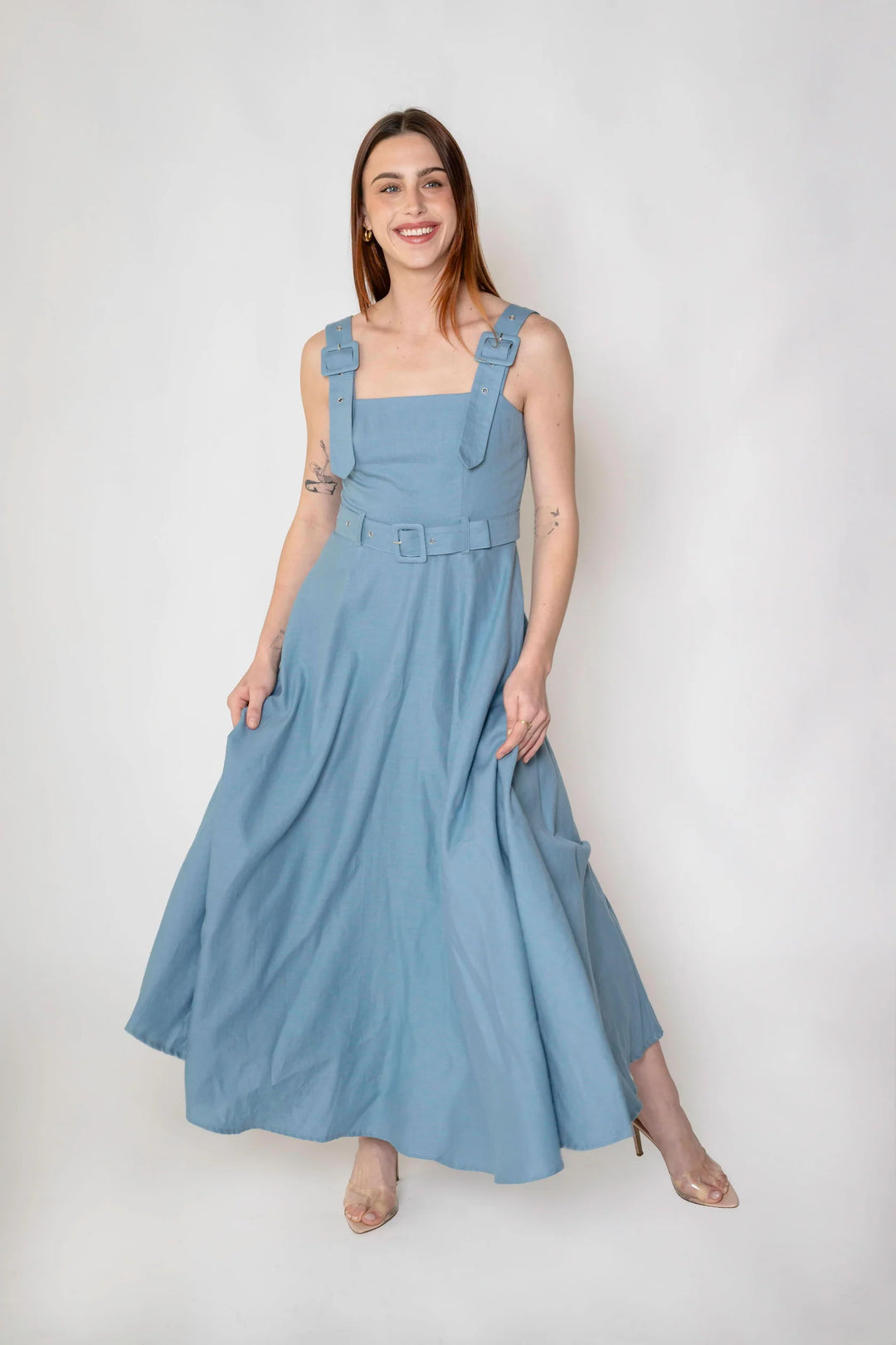 A woman modeling a dusty blue linen maxi dress with shoulder straps, a matching belt, and side pockets, standing against a light gray studio background.